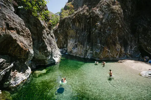 A small group of people, some with pool floats are shown in the clear, green-tinged water, with a small sand beach and cliffs behind them. 